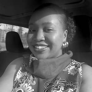 A greyscale headshot of Negi Chemaiywa, a Black woman wearing a patterned top, a scarf, and multiple earrings. She's smiling at the camera.