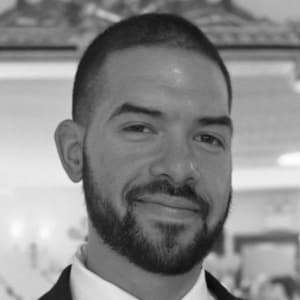 A black and white portrait of Jose Nieves-Santiago, a man with dark, close-cropped hair and a dark, close-cropped full beard and goatee. He is wearing a white collared shirt and a dark blazer.