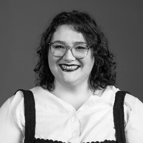 Kristen Corless, a light-skinned person with dark, curly shoulder-length hair. They are wearing a white collared shirt and glasses.